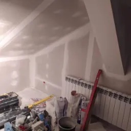 Poddasze w trakcie wykańczania, ściany pokryte płytami gipsowo-kartonowymi, widoczne worki z gipsem, narzędzia i grzejnik.