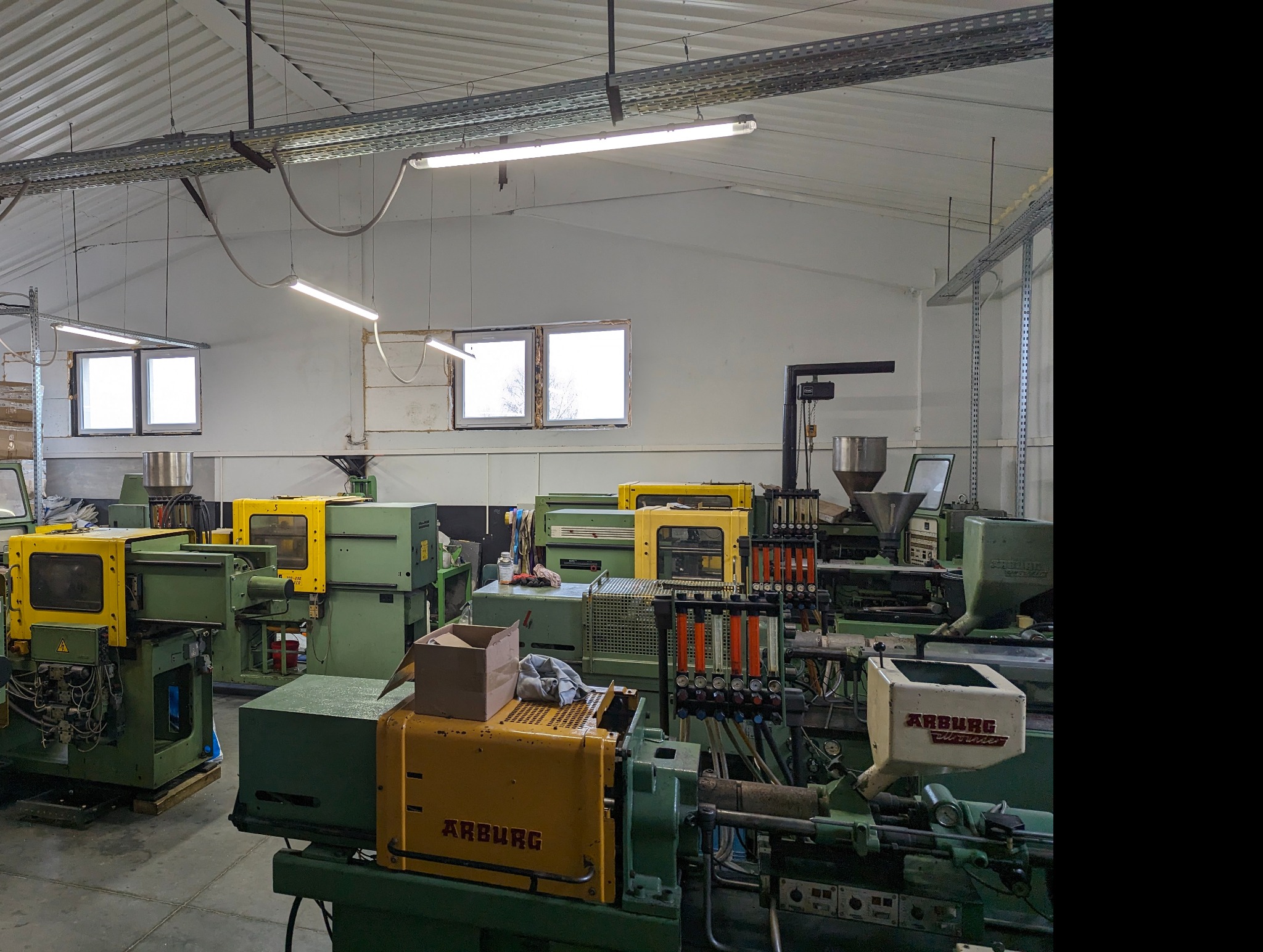 Wnętrze hali produkcyjnej z kilkoma maszynami w odcieniach zieleni i żółci, prawdopodobnie w trakcie pracy, oświetlone jarzeniówkami, widoczne okna i instalacje pod sufitem.