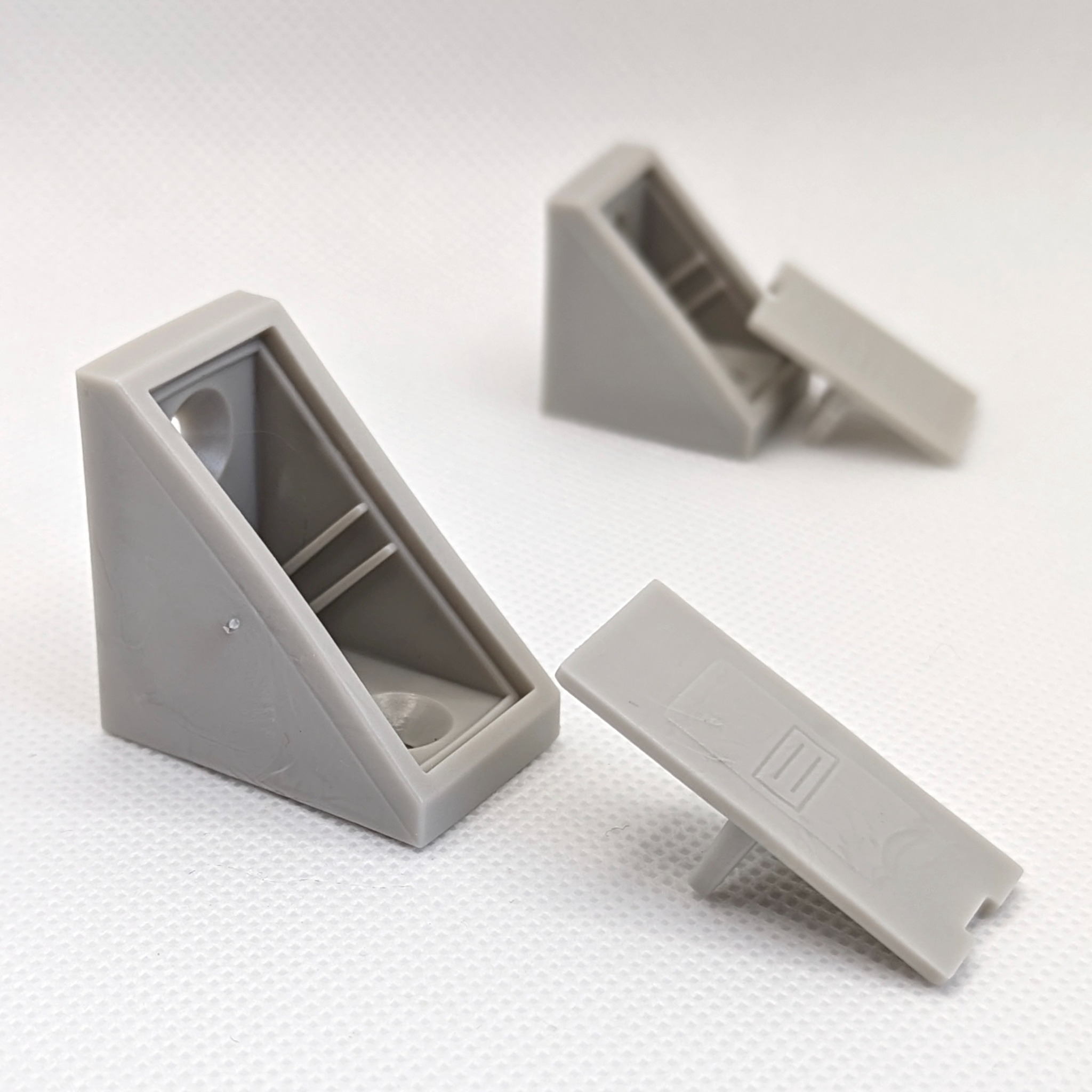 Dwa szare, plastikowe kątowniki z zaślepkami na białym tle. Jeden kątownik z zaślepką włożoną, drugi z zaślepką obok.
