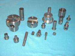 Zestaw precyzyjnie obrobionych, metalowych elementów o różnych kształtach i rozmiarach, leżących na jasnoniebieskim tle. Widoczne są walce, tuleje, dyski z rowkami i otwory, sugerujące zastosowanie...