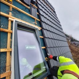 Uaługi Blacharsko-Dekarskie ART-Dachy - Montaż okna dachowego FAKRO na dachu pokrytym szarą dachówką, widoczne narzędzia i dekarz w odblaskowej kurtce podczas pracy.
