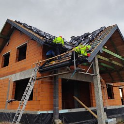 Uaługi Blacharsko-Dekarskie ART-Dachy - Ekipa dekarzy w jaskrawych ubraniach roboczych montuje dachówkę na nowym, ceglanym domu w stanie surowym otwartym, widoczna więźba dachowa i rusztowania.