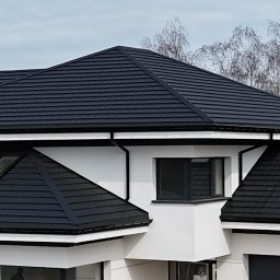 Uaługi Blacharsko-Dekarskie ART-Dachy - Nowoczesny dom z ciemną dachówką modułową, widoczne okna dachowe, rynny w kolorze dachu, biała elewacja i drzewa w tle.