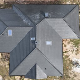 Uaługi Blacharsko-Dekarskie ART-Dachy - Widok z góry na nowy, grafitowy dach domu jednorodzinnego z oknami dachowymi i kominem, częściowo jeszcze mokry po deszczu, otoczony piaszczystym terenem.