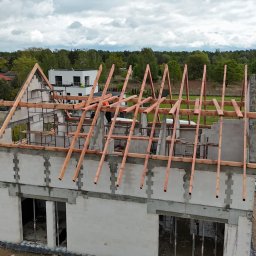 Uaługi Blacharsko-Dekarskie ART-Dachy - Wznoszenie drewnianej więźby dachowej na surowym budynku z betonu komórkowego. W tle zabudowa jednorodzinna i zieleń.