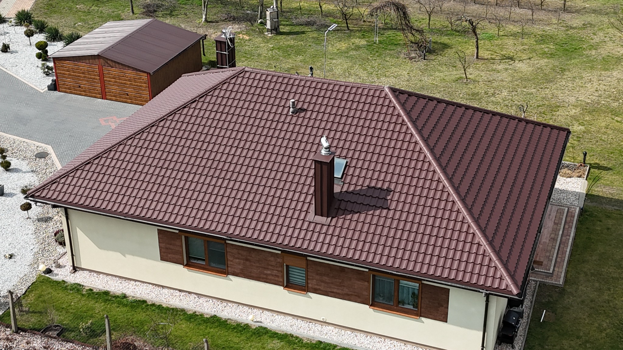 Widok z góry na dom z nowym, brązowym dachem z blachodachówki, kominem i wentylacją, w tle budynek gospodarczy z dachem w tym samym kolorze