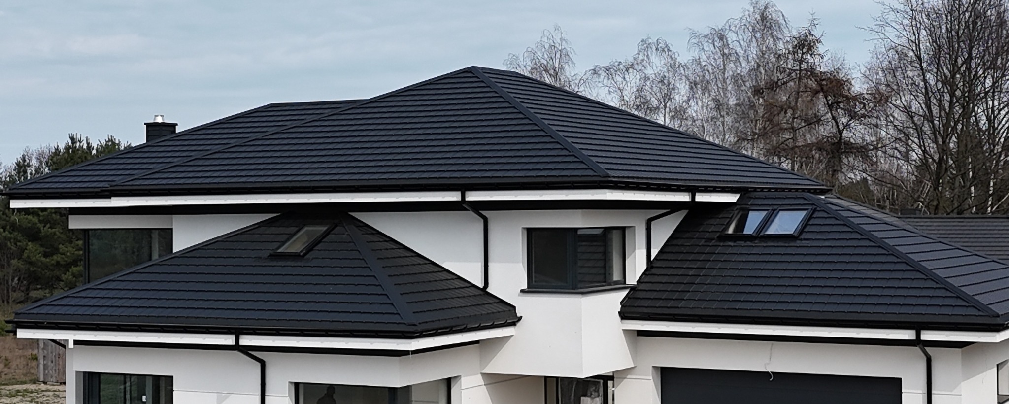 Nowoczesny dom z ciemną dachówką modułową, widoczne okna dachowe, rynny w kolorze dachu, biała elewacja i drzewa w tle.