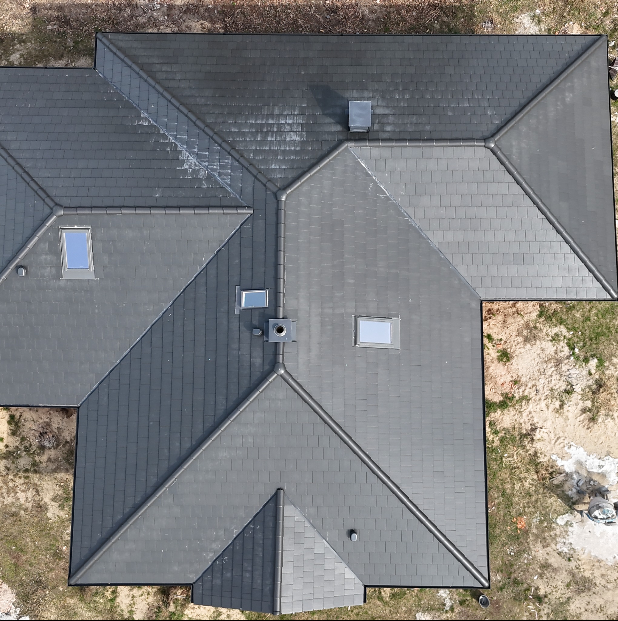 Widok z góry na nowy, grafitowy dach domu jednorodzinnego z oknami dachowymi i kominem, częściowo jeszcze mokry po deszczu, otoczony piaszczystym terenem.