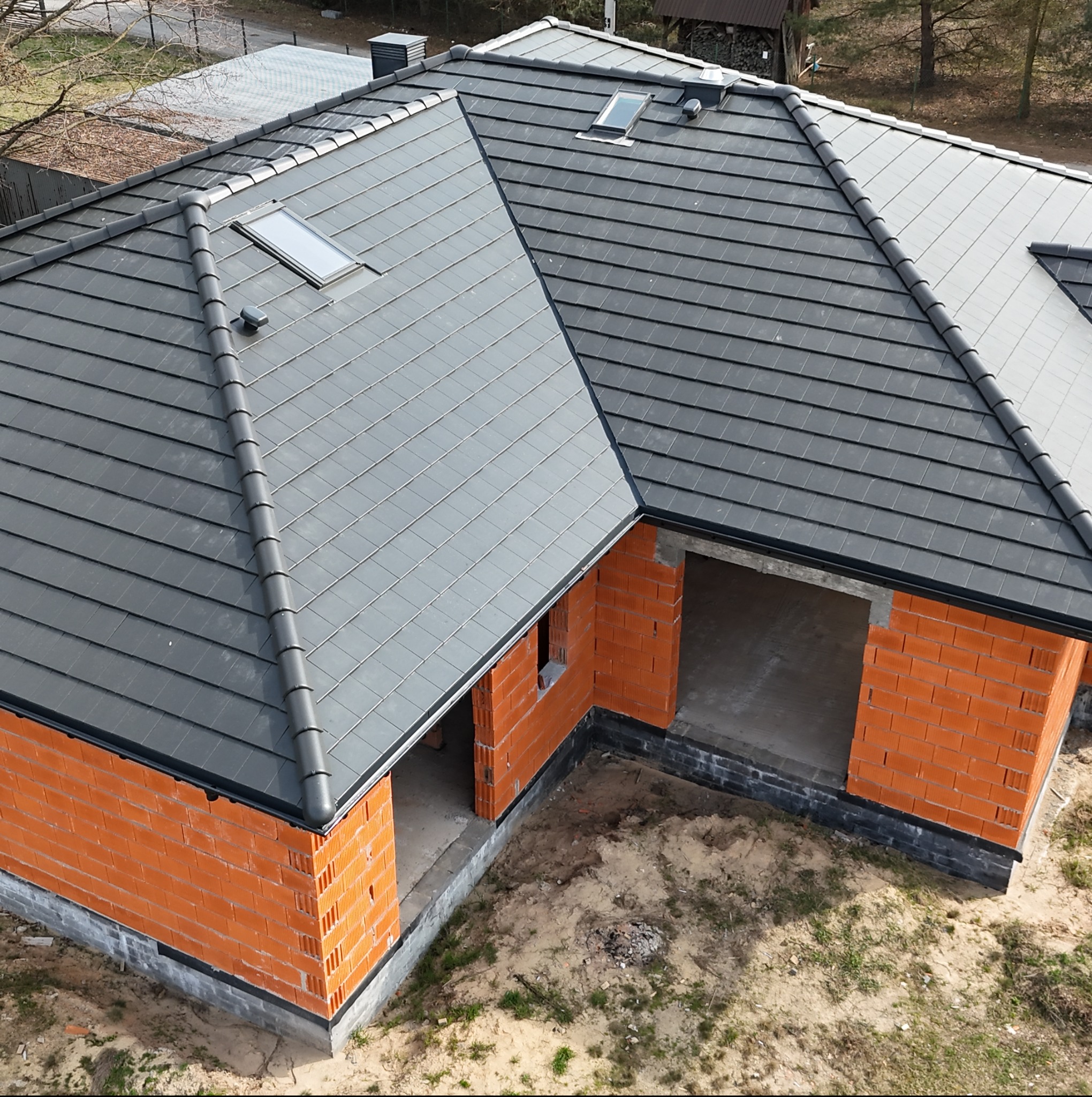 Nowy, grafitowy dach z dachówki ceramicznej z oknami dachowymi na budynku w stanie surowym otwartym z czerwonej cegły.
