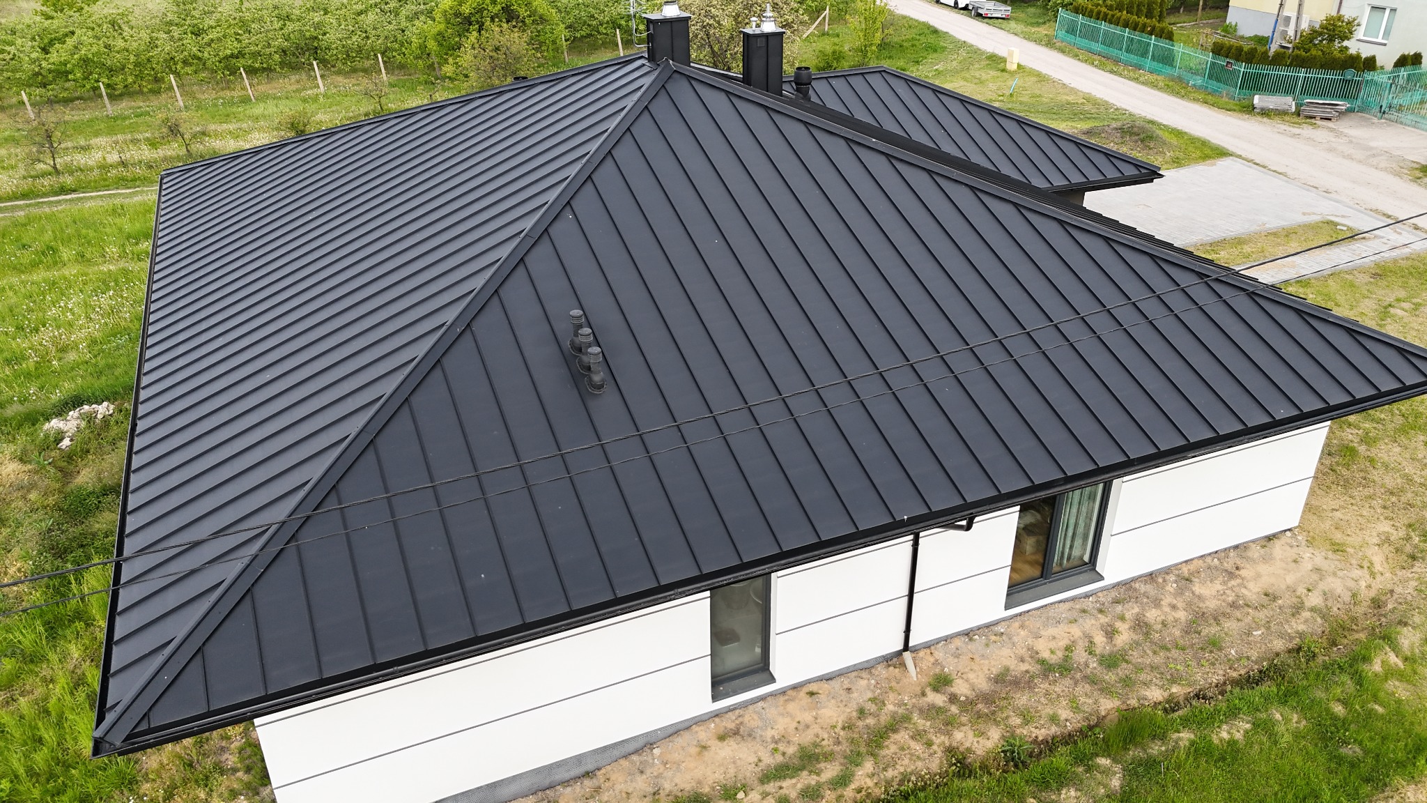 Czarny dach z blachy na nowoczesnym domu z białymi ścianami, widok z góry, kominy i elementy wentylacyjne na dachu.