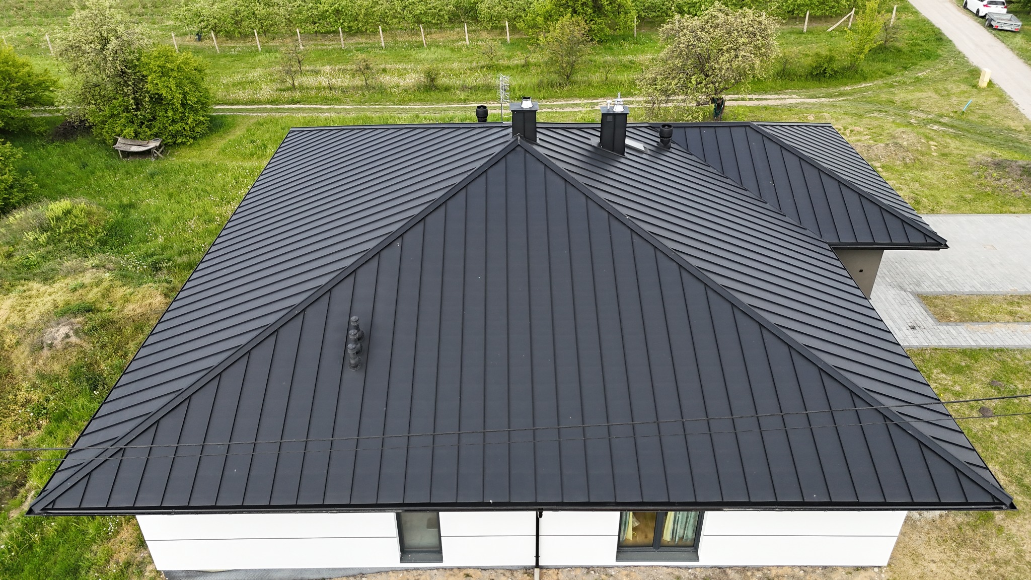 Czarny, blaszany dach domu jednorodzinnego z kominami, widziany z góry na tle zieleni i zabudowy. Nowoczesne pokrycie dachowe z wyraźnymi liniami.