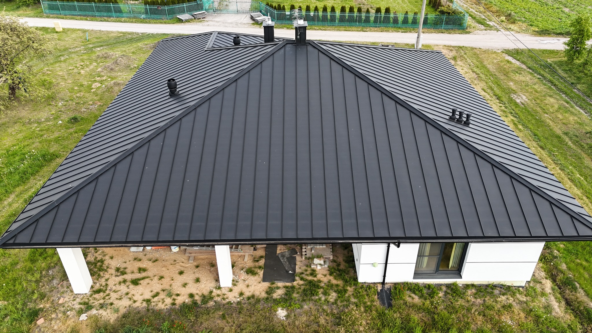 Widok z góry na nowoczesny dom z ciemnoszarym dachem z blachy na rąbek stojący. Widoczne kominy i lukarny. Dom otoczony zielenią, w tle ogrodzenie.