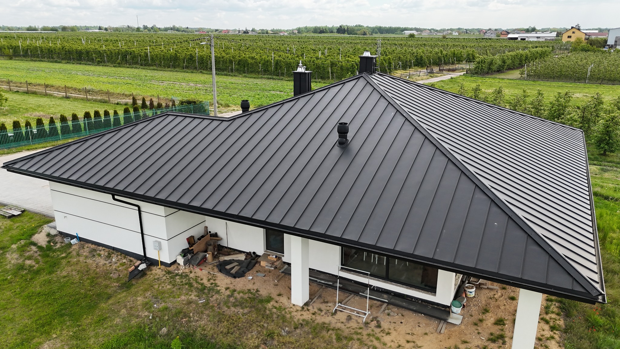 Nowoczesny dom z czarnym, metalowym dachem na tle sadu. Widok z góry ukazuje geometrię dachu i minimalistyczną architekturę budynku. Detale: kominy, rynny.