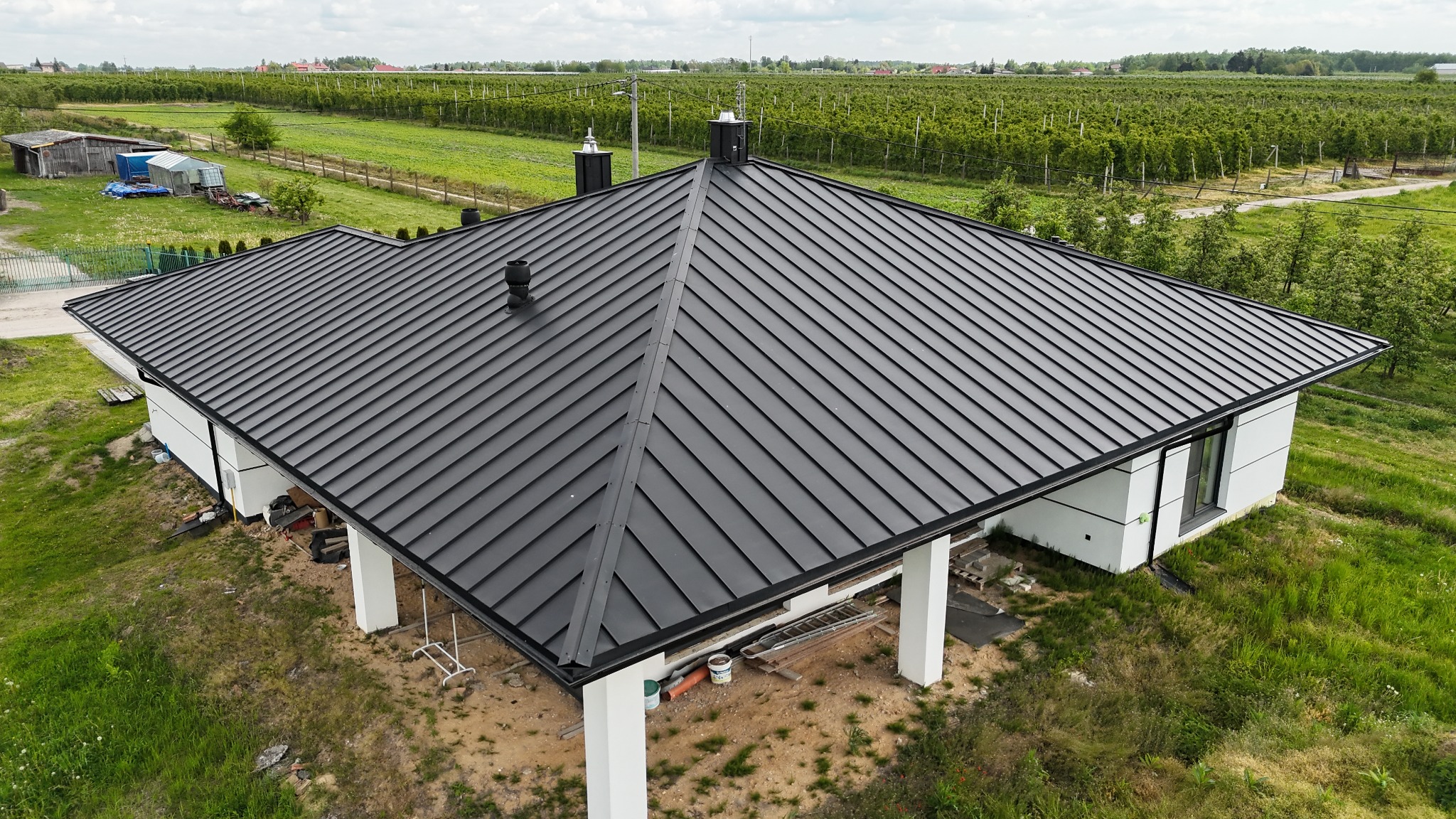 Czarny, blaszany dach domu jednorodzinnego z kominami, widziany z góry na tle zieleni i sadu. Widoczne detale wykończenia i otoczenie budynku.