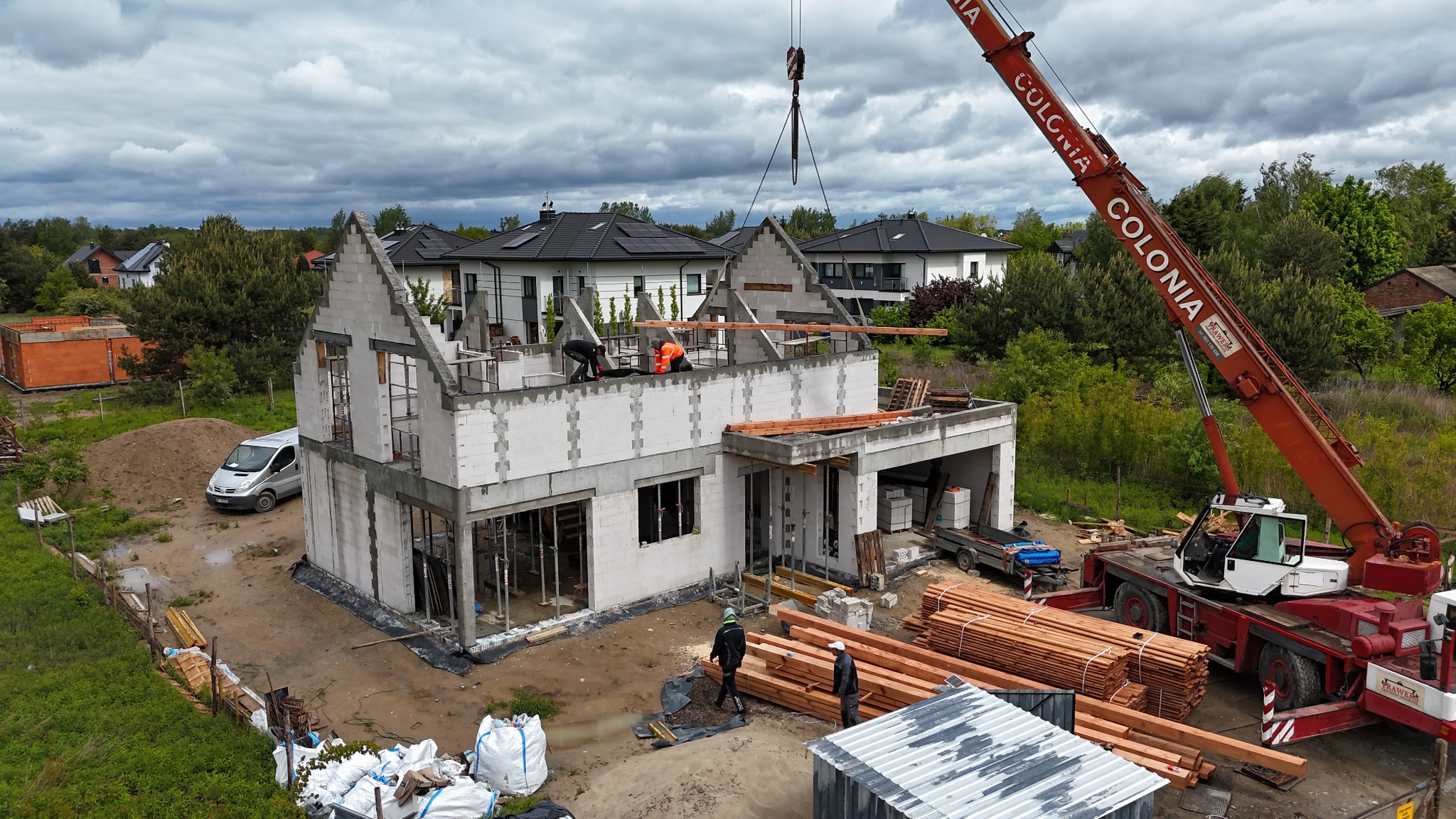 Budowa domu jednorodzinnego w stanie surowym otwartym z użyciem dźwigu, widoczne elementy konstrukcyjne i pracownicy w trakcie montażu więźby dachowej.