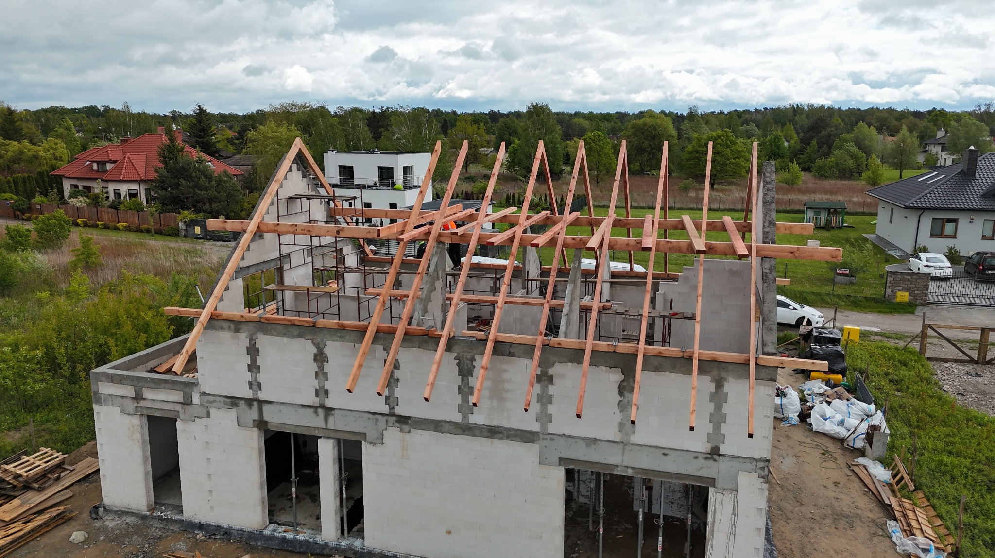 Wznoszenie drewnianej więźby dachowej na surowym budynku z betonu komórkowego. W tle zabudowa jednorodzinna i zieleń.