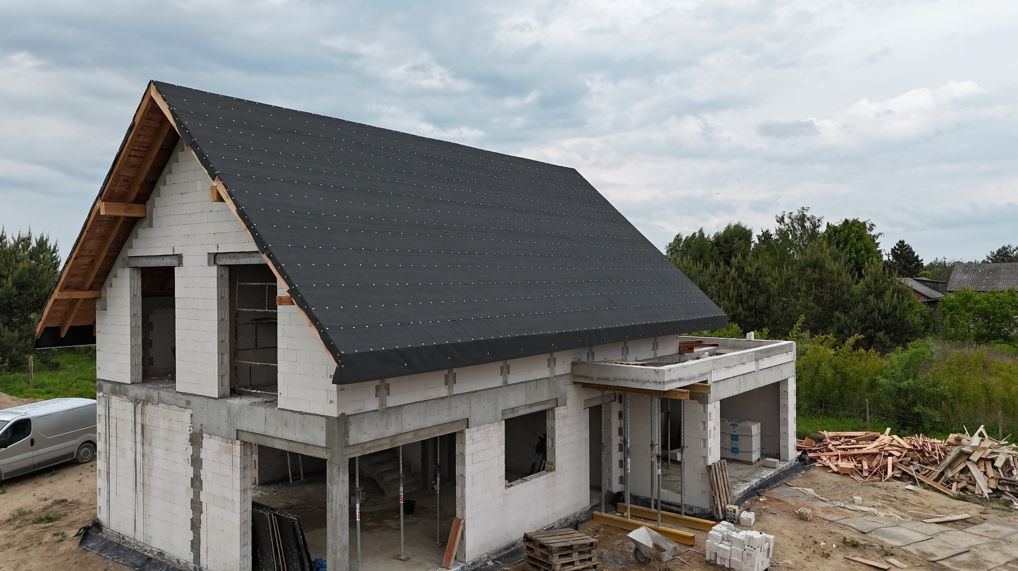 Dom w trakcie budowy z czarną membraną dachową, widoczne pustaki, drewniana więźba i materiały budowlane. W tle zieleń i pochmurne niebo.