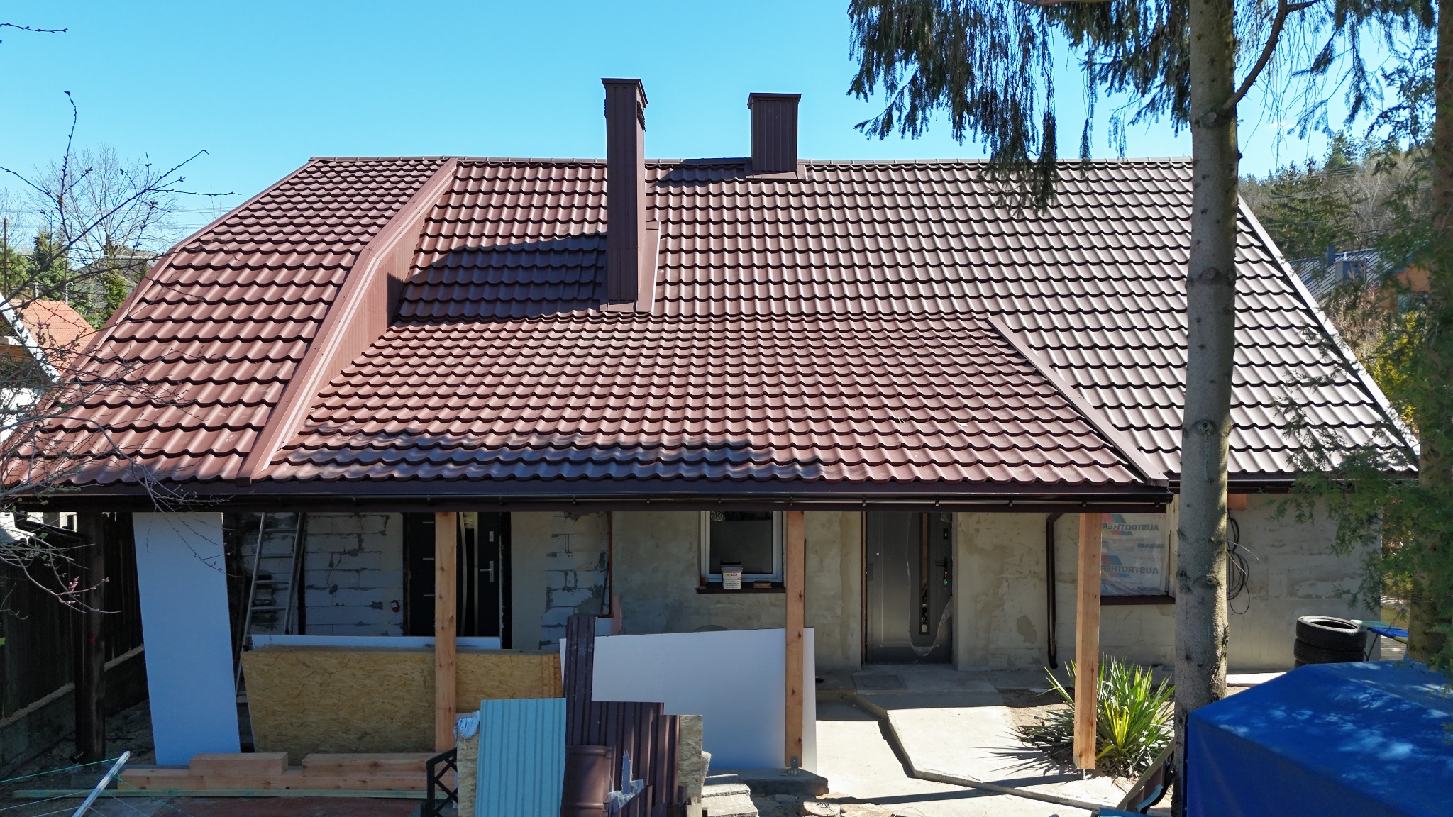 Brązowy dach z blachodachówki na parterowym budynku z dwoma kominami, częściowo w trakcie budowy, z widocznymi materiałami budowlanymi.