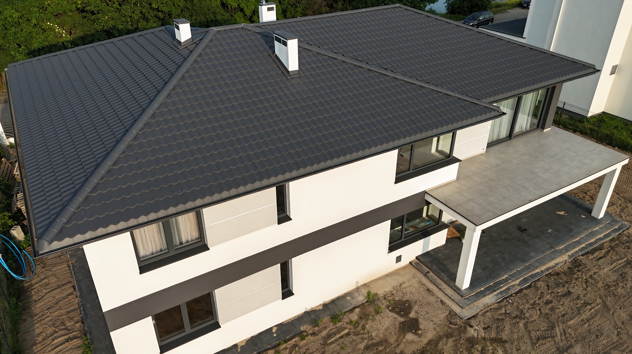 Nowoczesny dom z grafitowym dachem z dachówki ceramicznej, widok z góry. Biała elewacja z szarymi akcentami, duży taras. Elementy wykończeniowe.