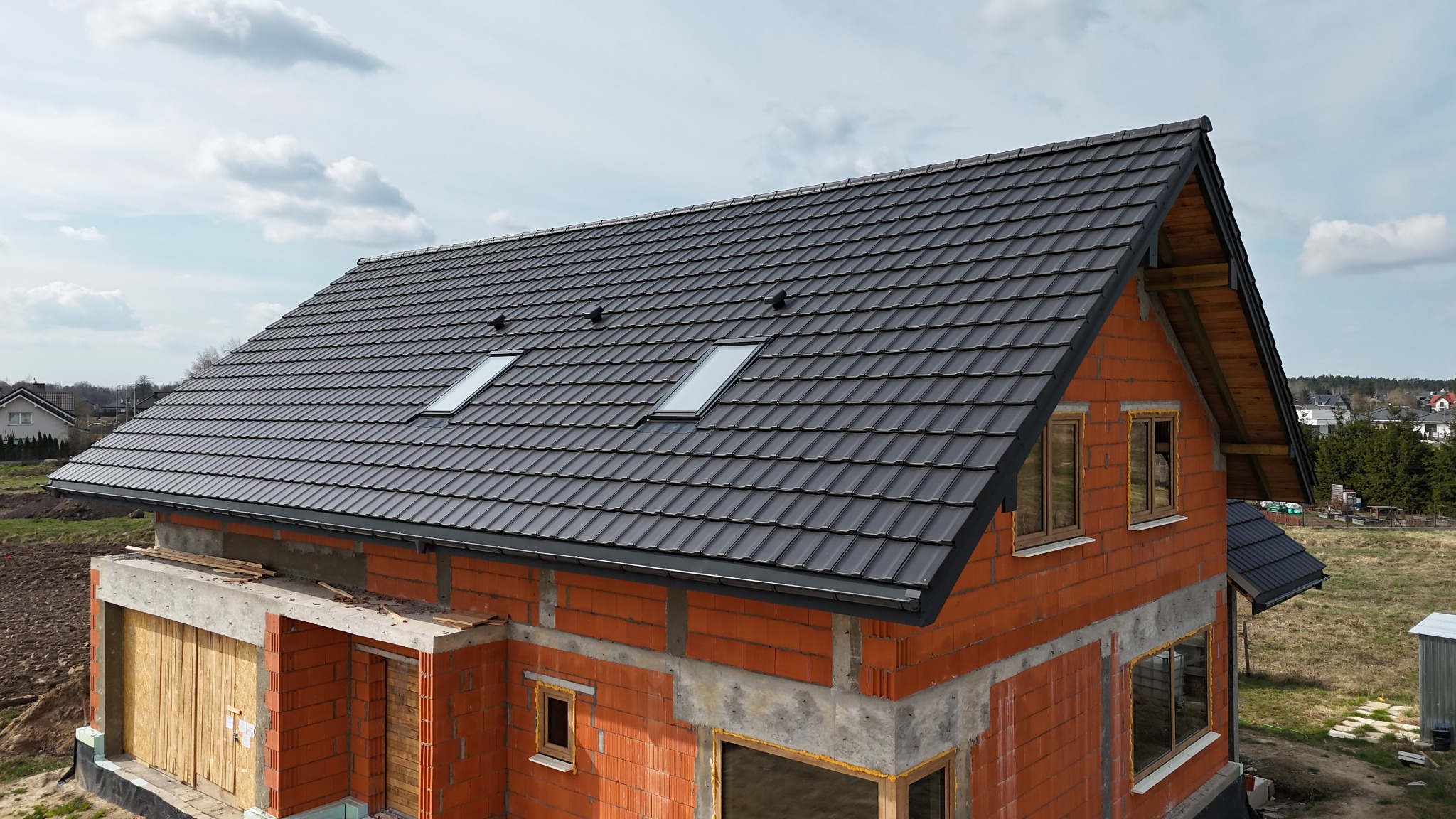 Dom w trakcie budowy z ciemnoszarym dachem z dachówki ceramicznej i oknami dachowymi, widok z góry, częściowo otynkowane ściany z cegły.