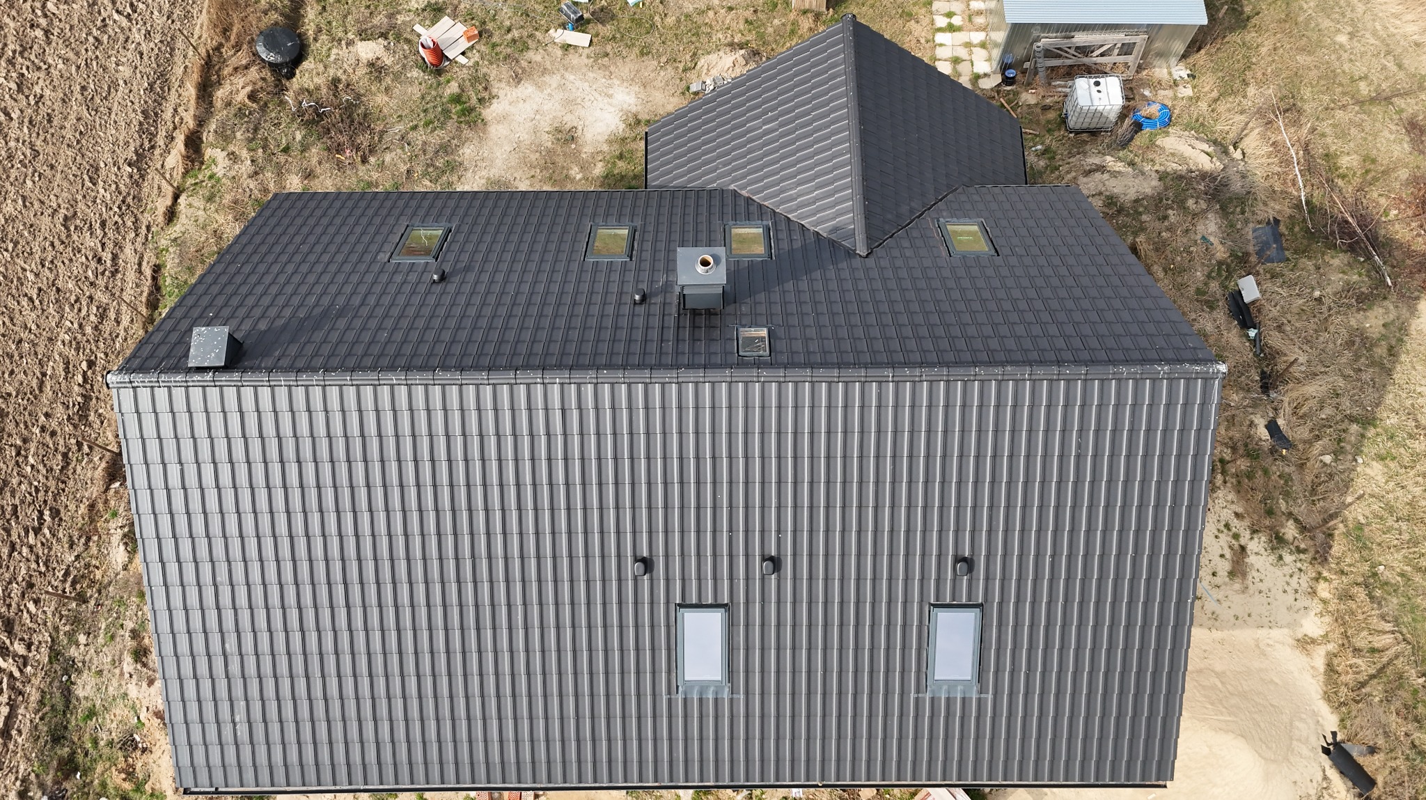 Widok z góry na nowy, ciemnoszary dach z dachówki ceramicznej. Widoczne okna dachowe, komin wentylacyjny i obróbki blacharskie. W tle zabudowania gospodarcze i niezagospodarowany teren.