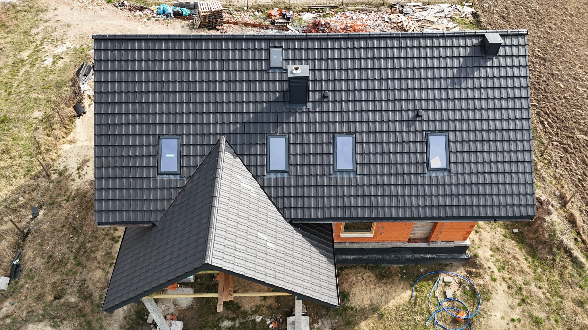 Widok z góry na dom w trakcie budowy z ciemnoszarą dachówką, widoczne okna dachowe i komin. Część dachu jest już pokryta, a część budynku w stanie surowym.