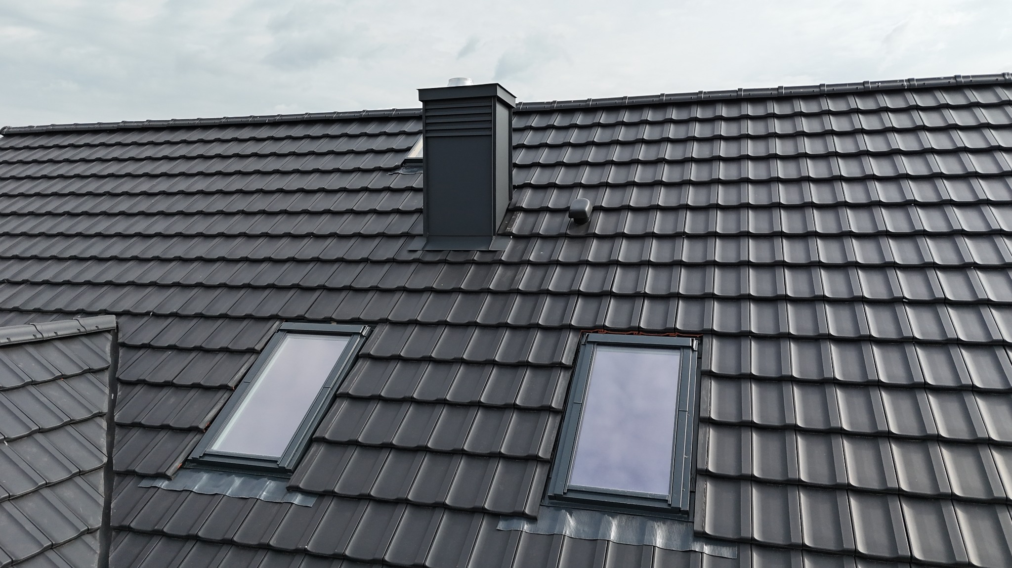 Grafitowy dach z dachówki ceramicznej z dwoma oknami połaciowymi i kominem, widok z góry. Nowoczesne pokrycie dachowe w stonowanej kolorystyce.