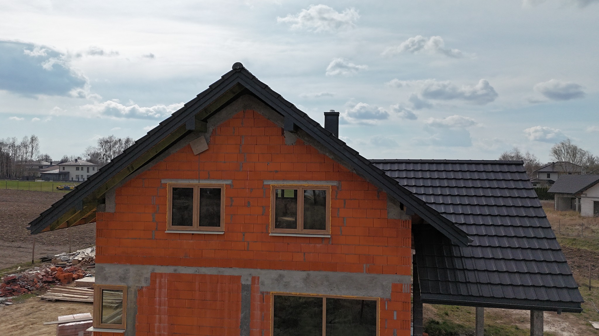 Dom w trakcie budowy: ściany z czerwonej cegły, fragment dachu z czarnej dachówki. Widoczne okna z drewnianą ramą i elementy konstrukcji dachu. Pochmurne niebo w tle.