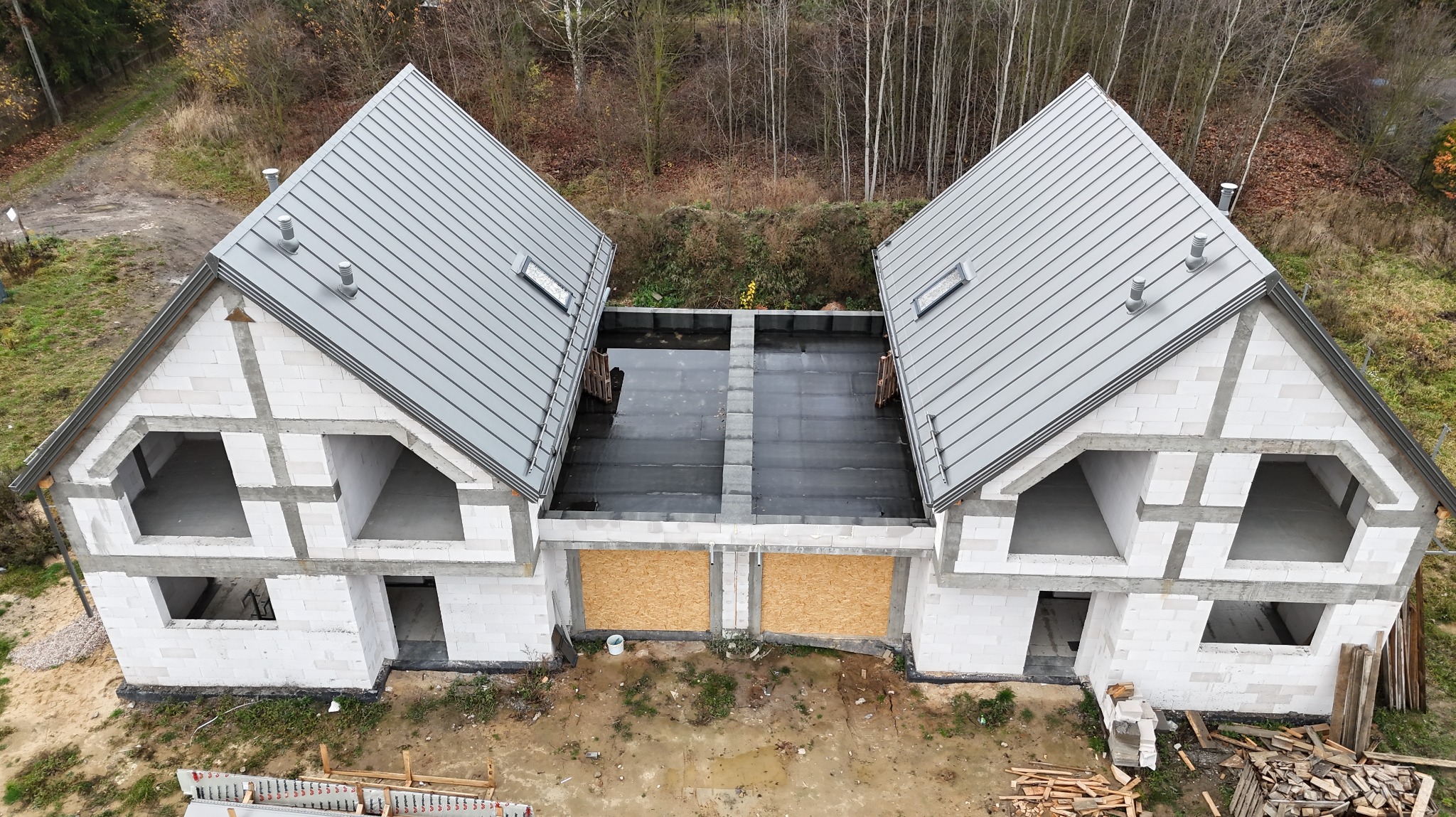 Dwa domy w stanie surowym zamkniętym z szarym dachem z blachy na tle drzew. Widok z góry ukazuje połączone dachy i zabezpieczone otwory okienne.
