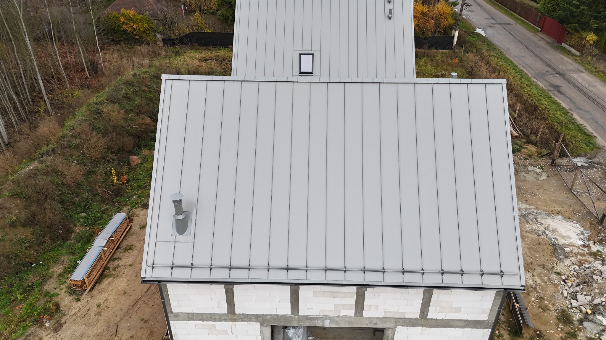Nowoczesny, szary dach z blachy na budynku w trakcie budowy, widok z góry. Na dachu komin wentylacyjny i okno dachowe. W tle drzewa i droga.