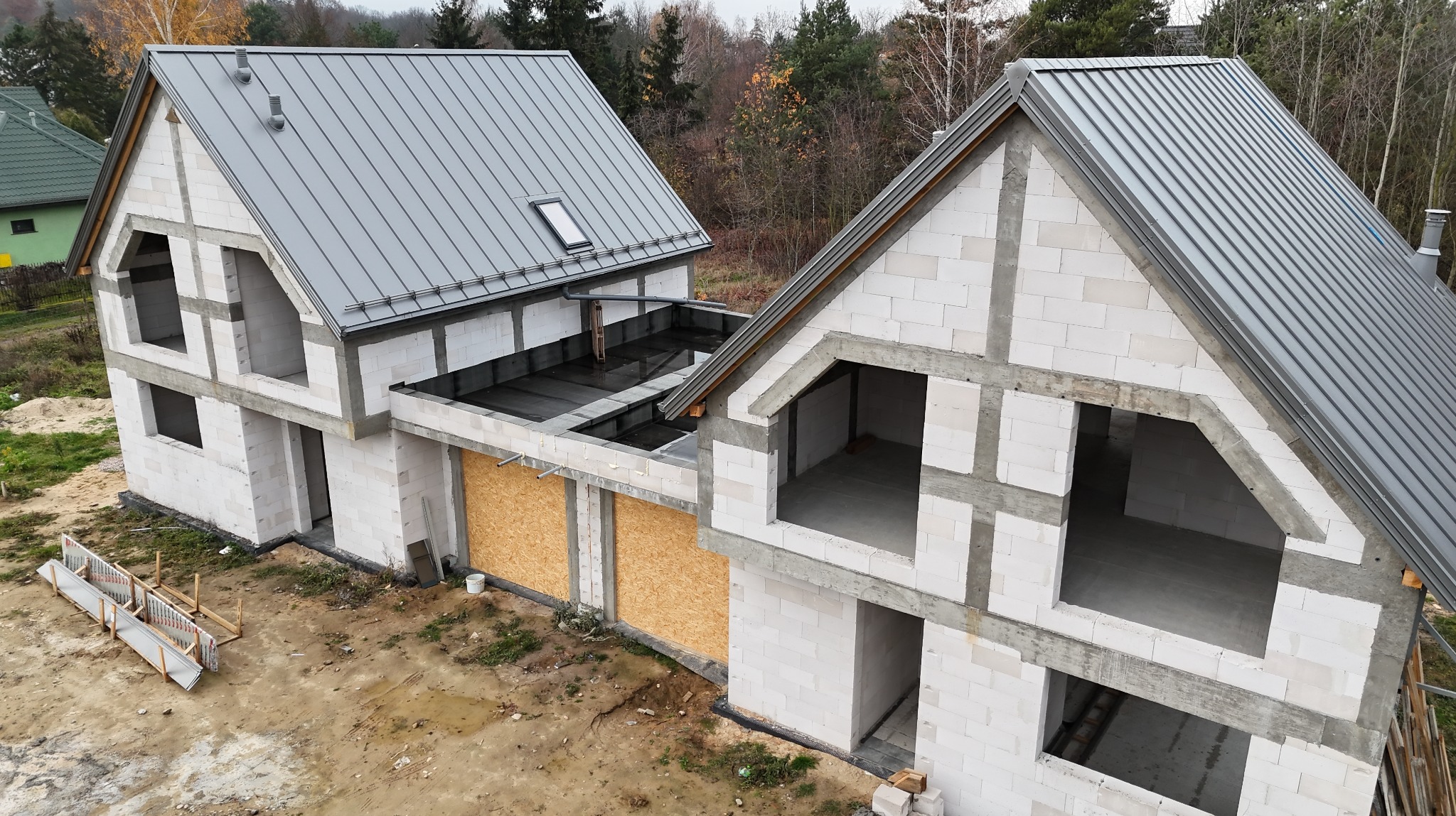 Surowy stan dwukondygnacyjnego domu z szarym dachem z blachy na tle drzew. Widoczne białe bloczki konstrukcyjne i okna dachowe. Częściowo zabite deskami.