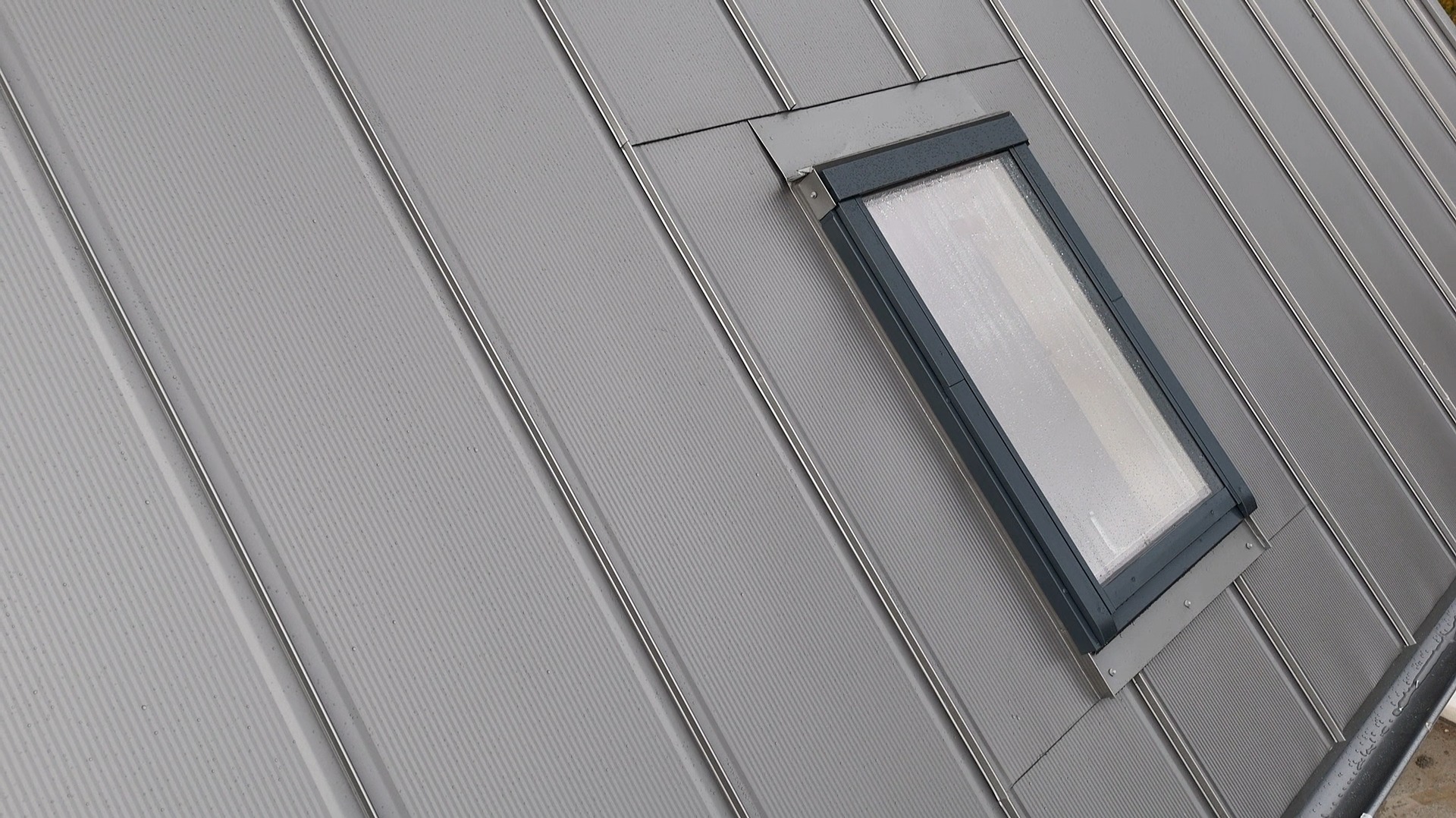 Szary dach z blachy na rąbek stojący z oknem dachowym. Widoczne detale łączeń blachy i obróbki okna. Na oknie widoczne krople deszczu.