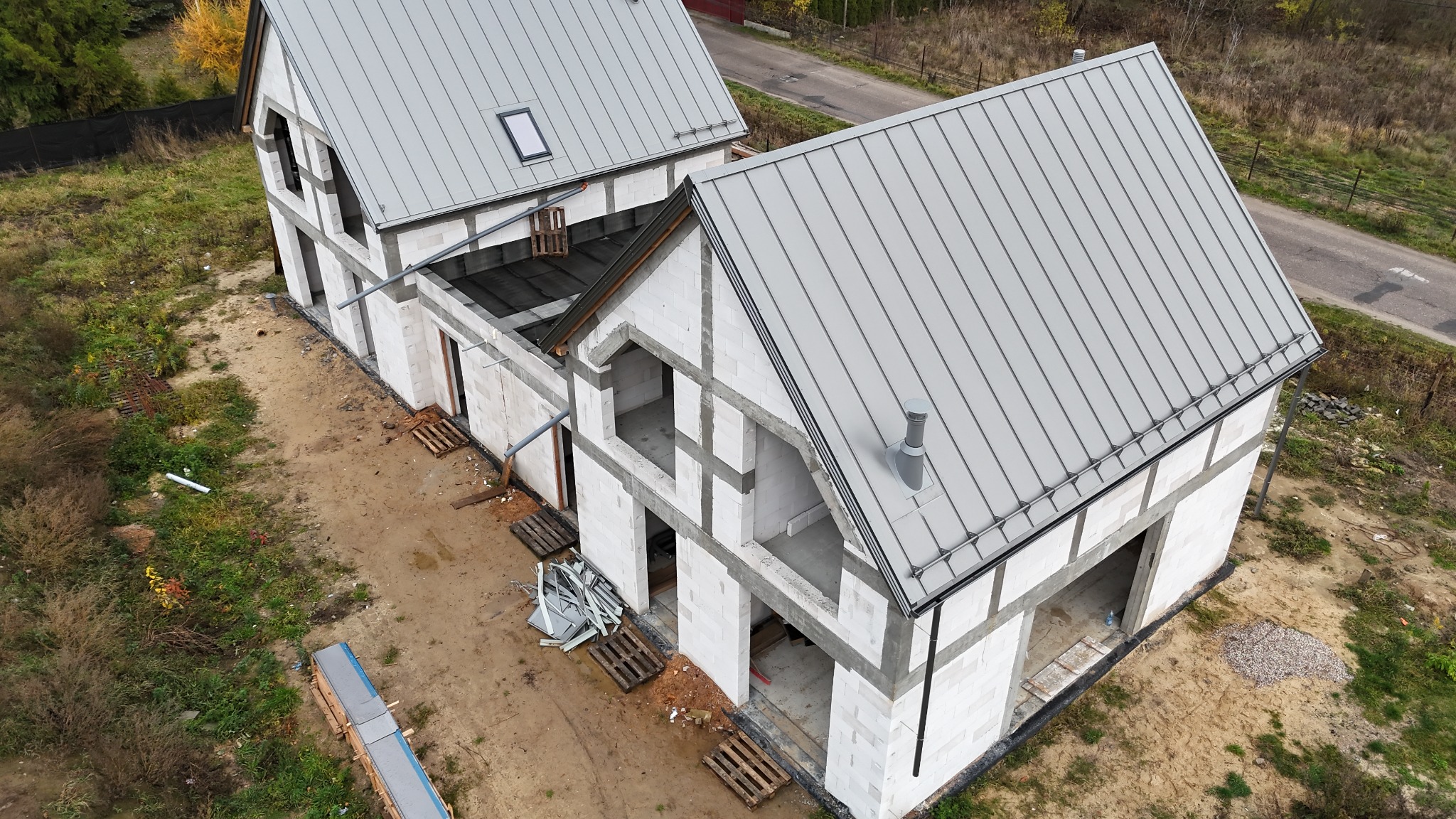 Wysoka perspektywa: Dwa budynki w trakcie budowy, jeden z szarym dachem z blachy na ukończeniu, drugi w stanie surowym, białe ściany z betonu komórkowego.