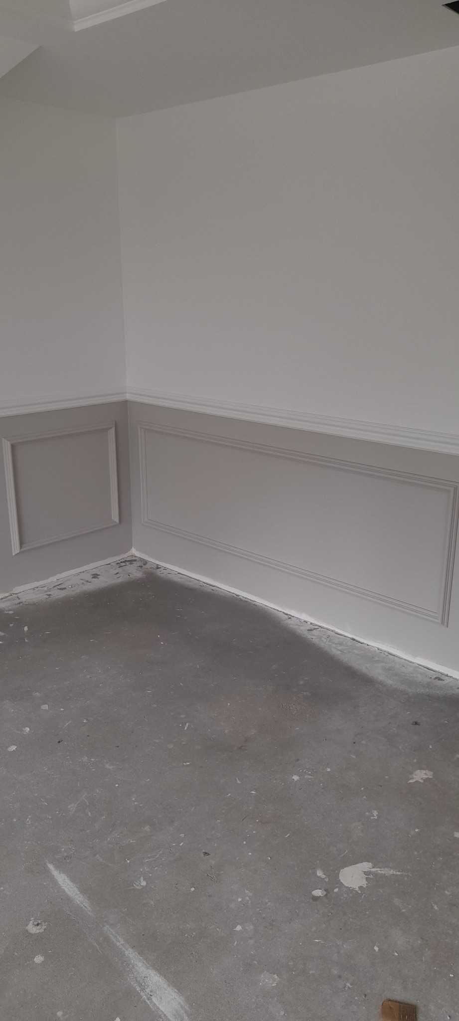 Świeżo pomalowane ściany z dekoracyjnymi panelami w dolnej części, szare panele i biała górna część ścian, surowa betonowa podłoga z plamami farby, narożnik pokoju.