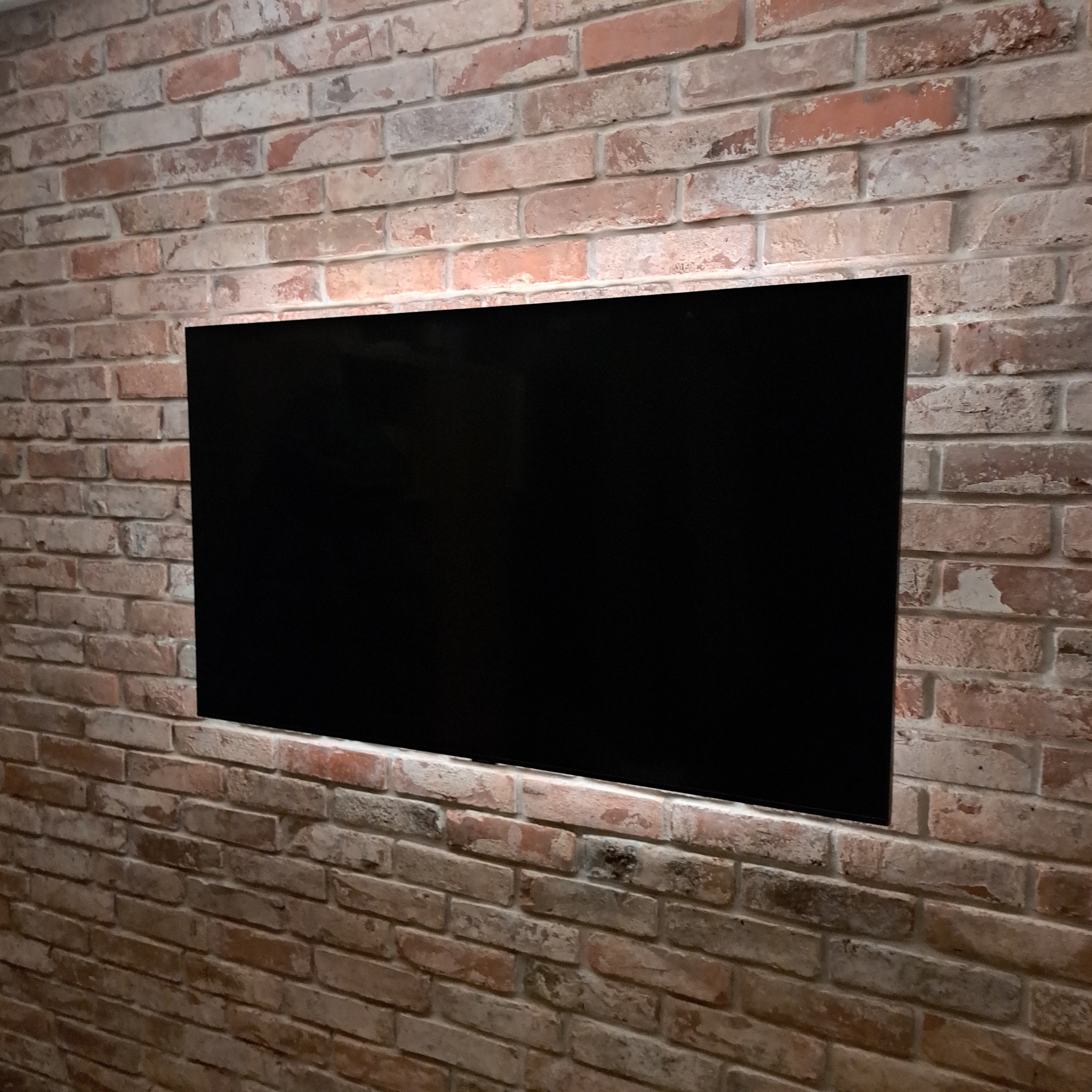 Telewizor z czarnym ekranem zamontowany na ścianie z cegły w stylu rustykalnym, widoczne nierówności i naturalne przebarwienia cegieł.