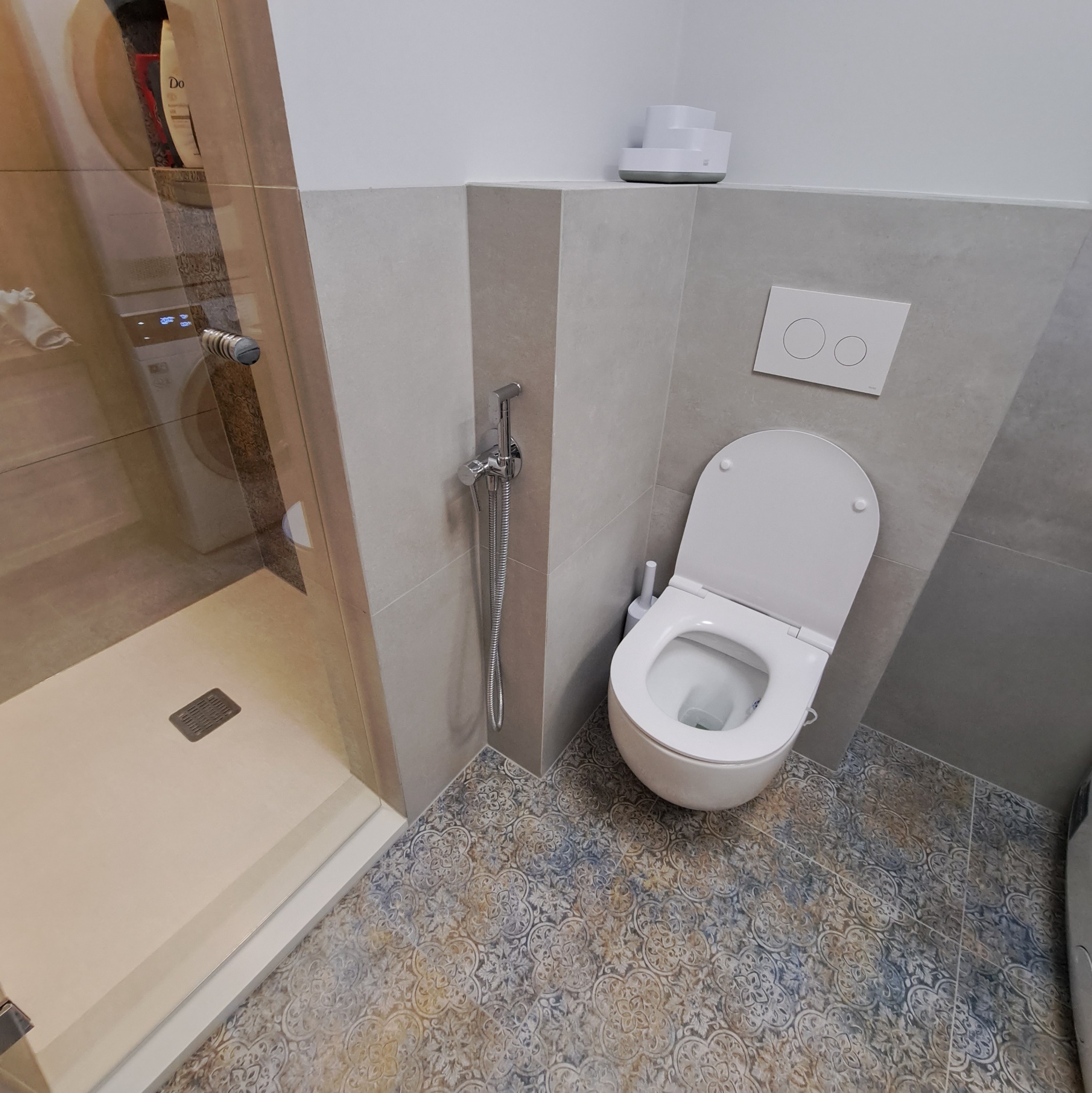 Nowoczesna łazienka po remoncie: biała toaleta wisząca, bidetka ze słuchawką, kabina prysznicowa ze szklanymi drzwiami, wzorzysta podłoga z płytek ceramicznych.