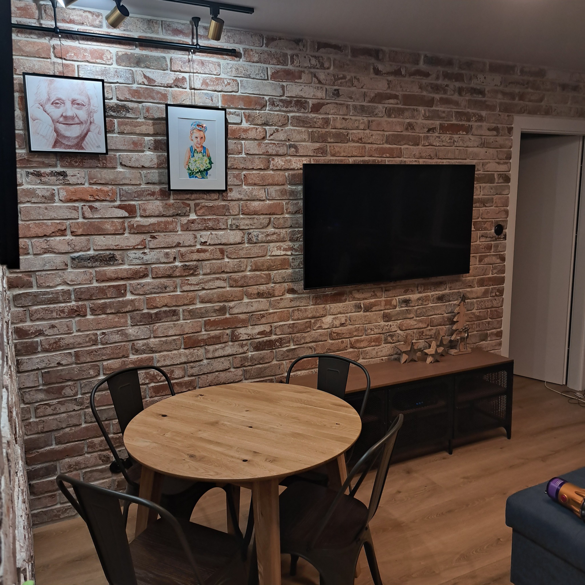 Salon z ceglaną ścianą, okrągłym drewnianym stołem i czarnymi krzesłami w stylu industrialnym, podświetlony reflektorami szynowymi, ozdobiony obrazami i telewizorem.