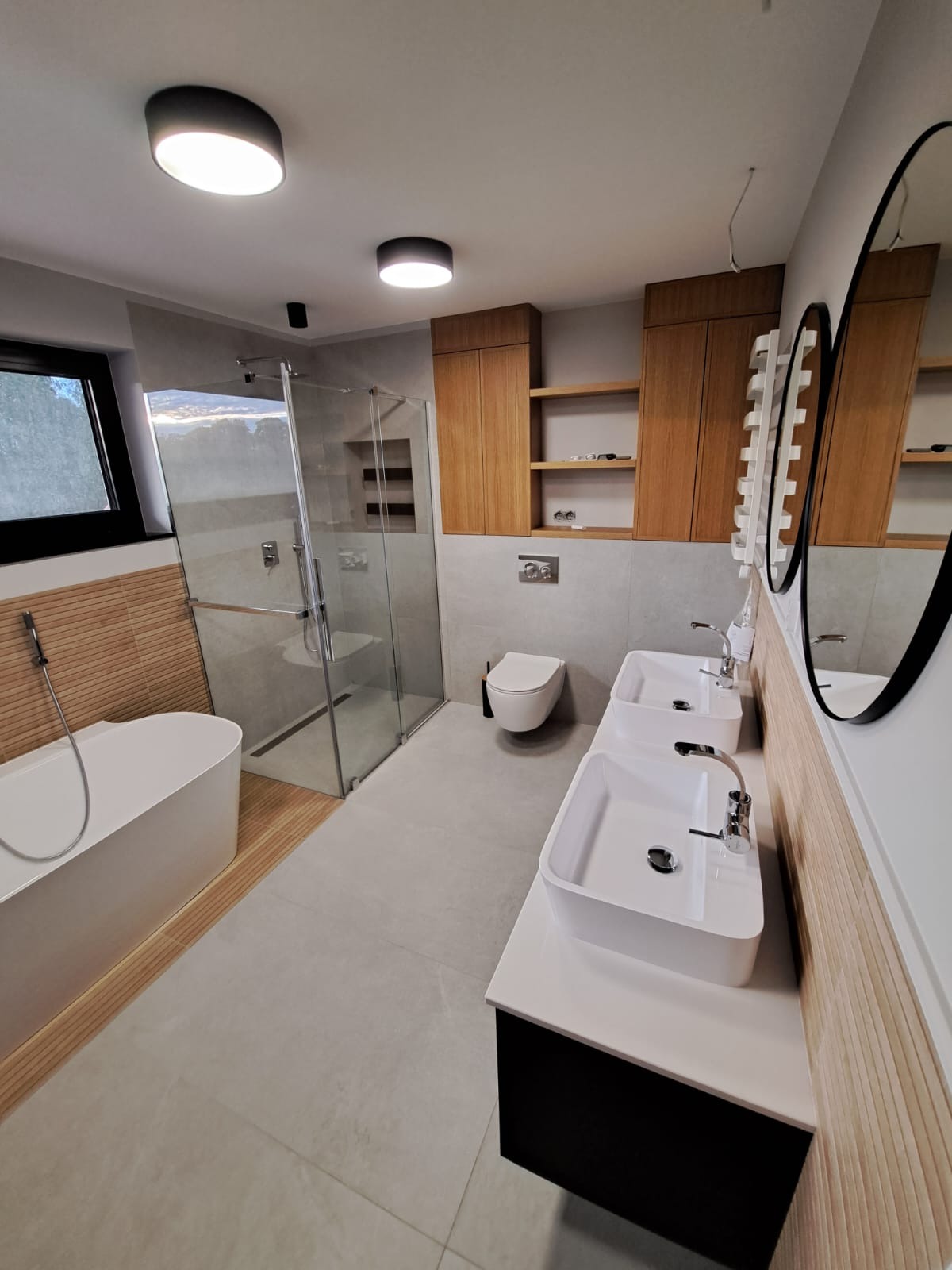 Nowoczesna łazienka z dwoma umywalkami na blacie, kabiną prysznicową typu walk-in, wolnostojącą wanną, drewnianymi szafkami wiszącymi i dekoracyjną ścianą z drewnopodobnych paneli.