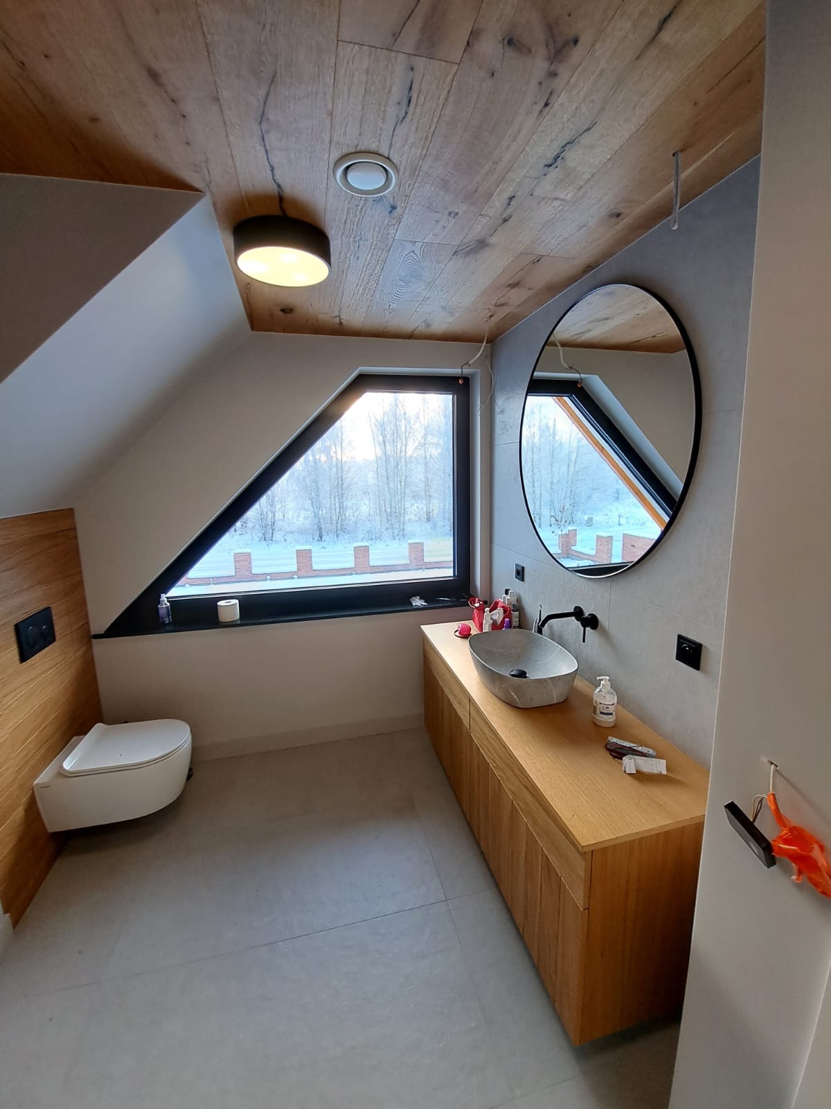 Nowoczesna łazienka na poddaszu z drewnianym sufitem i szafką pod umywalką, okrągłe lustro na szarej ścianie, widok na zimowy krajobraz za oknem o nietypowym kształcie.