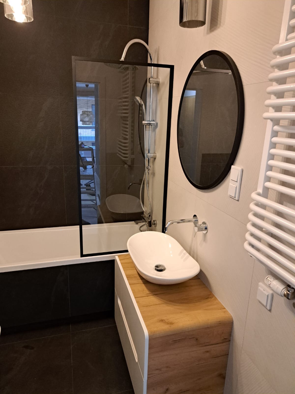 Nowoczesna łazienka z owalną umywalką nablatową na szafce z drewnianym blatem, czarną kabiną prysznicową typu walk-in i okrągłym lustrem w czarnej ramie.