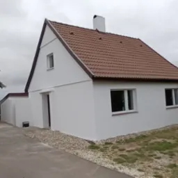 Biały dom z brązowym dachem po renowacji, widok z boku na tle zieleni i pochmurnego nieba.