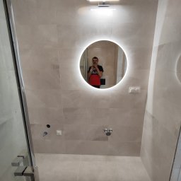 Usługi remontowo-budowlane Przemysław Felczak - Wykończona łazienka z beżowymi płytkami, okrągłym lustrem z podświetleniem LED i widocznymi przyłączami wodnymi oraz fragmentem kabiny prysznicowej.