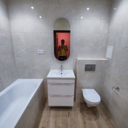 Usługi remontowo-budowlane Przemysław Felczak - Wykończona łazienka z beżowymi płytkami imitującymi marmur, drewnopodobną podłogą, białą wanną, umywalką z szafką i toaletą podwieszaną. W lustrze odbija się osoba robiąca zdjęcie.