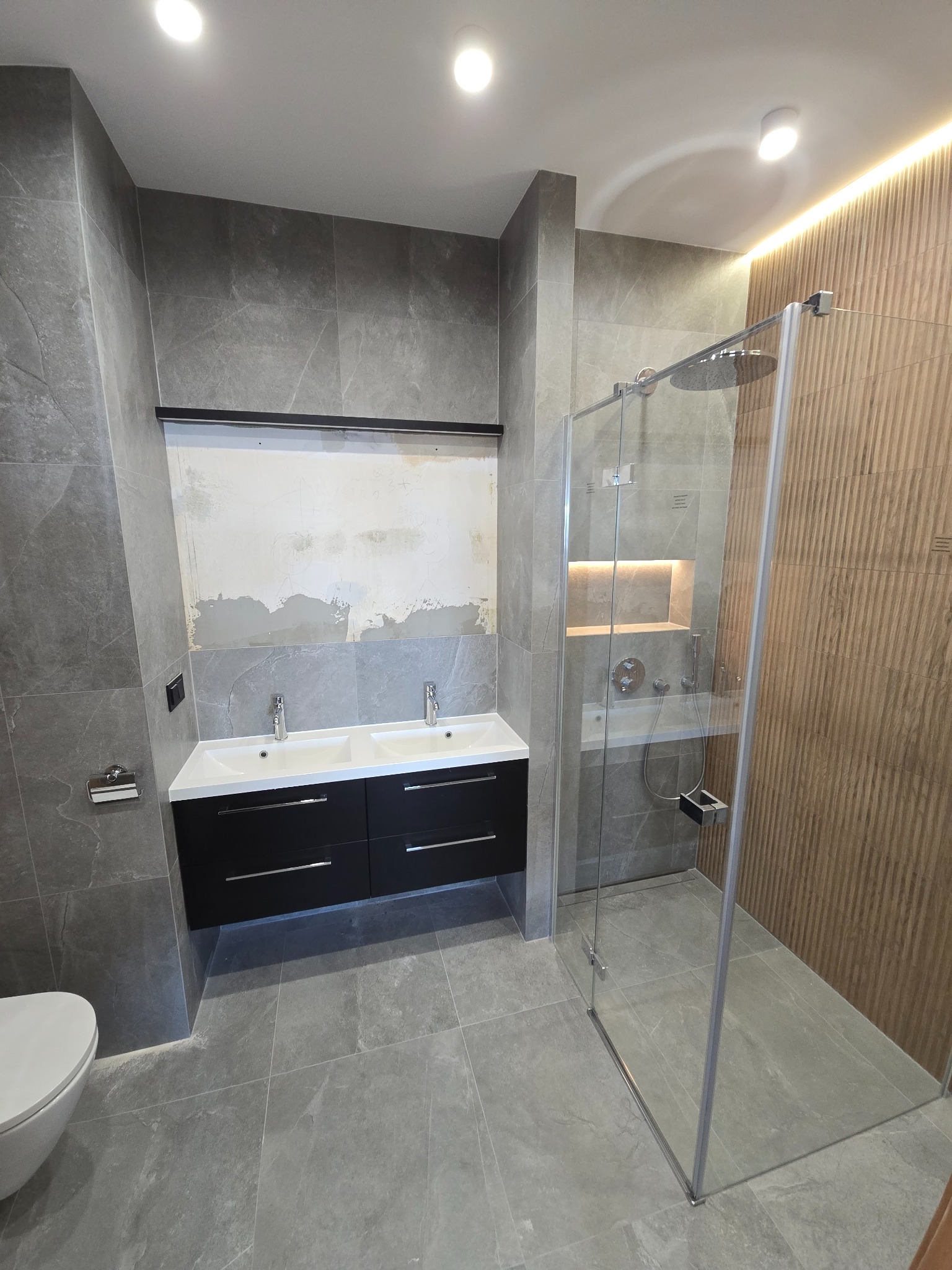 Nowoczesna łazienka w szarościach z kabiną prysznicową typu walk-in, podwójną umywalką z czarną szafką i częściowo wykończoną ścianą nad nią. Oświetlenie LED.