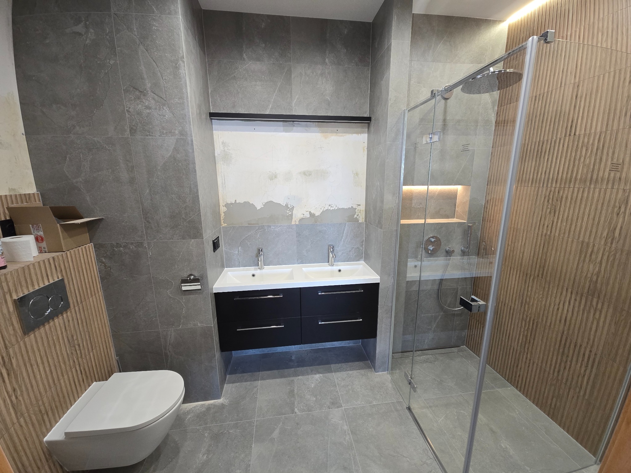 Nowoczesna łazienka w szarościach z kabiną prysznicową typu walk-in, podwójną umywalką i wiszącą toaletą. Częściowo wykończona ściana nad umywalką.