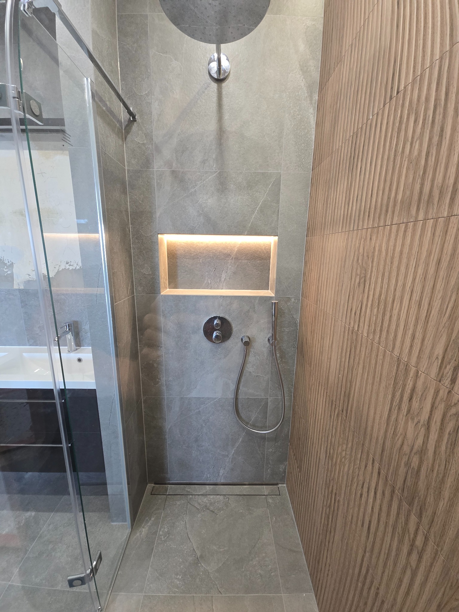 Nowoczesna kabina prysznicowa z szarymi płytkami imitującymi marmur i drewnopodobnymi płytkami na ścianie. Wnęka z podświetleniem. Minimalistyczny design.