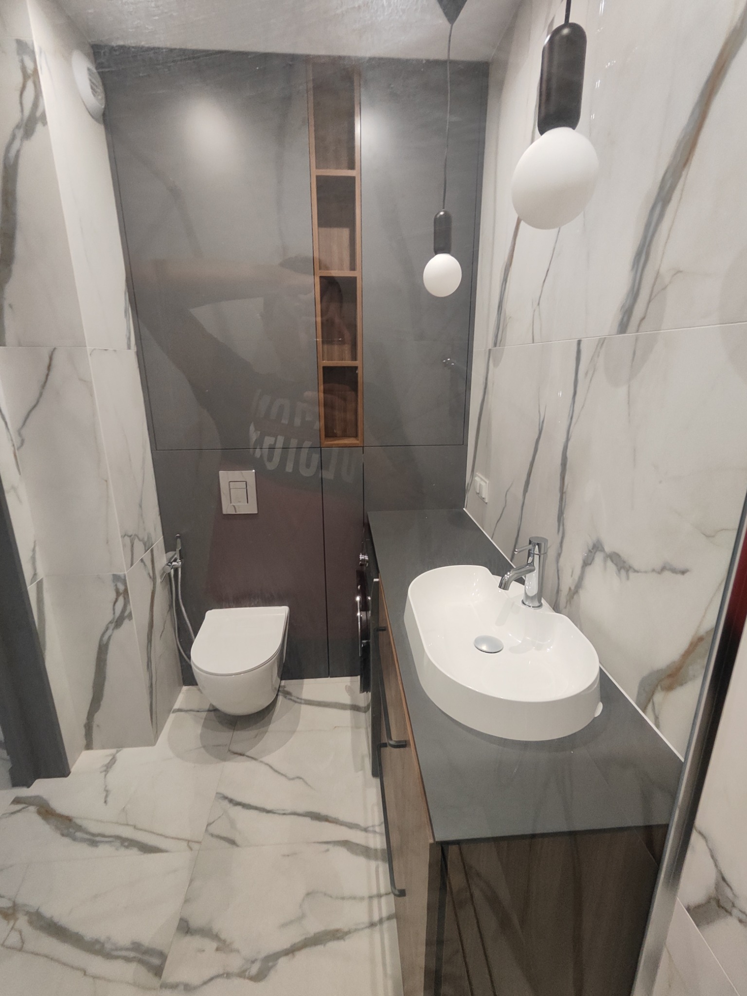 Nowoczesna łazienka w szaro-białych marmurkowych płytkach z wiszącą toaletą, owalną umywalką nablatową i minimalistycznym oświetleniem punktowym.