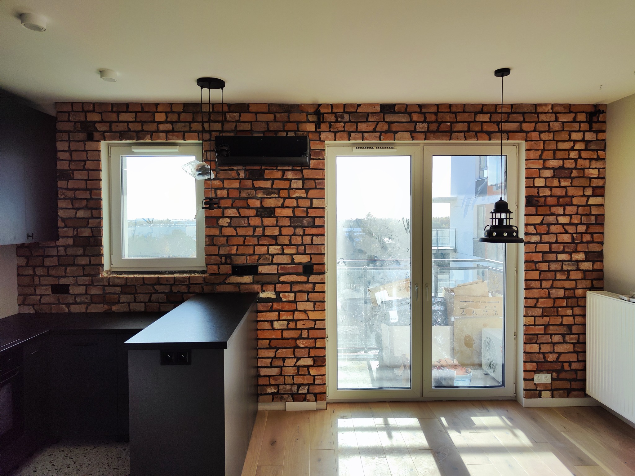 Kuchnia z ceglaną ścianą, czarnymi szafkami i oświetleniem, widok na balkon z klimatyzatorem.