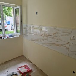 Oko - Fragment ściany w trakcie układania płytek imitujących marmur z widocznymi gniazdkami elektrycznymi, okno z widokiem na zewnątrz i nieporządek z narzędziami na podłodze.