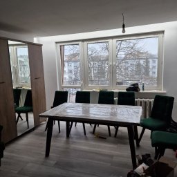 Oko - Pokój po remoncie z nową podłogą, stołem zabezpieczonym folią i zielonymi krzesłami. Widoczna szafa z lustrem i okno z widokiem na budynki.